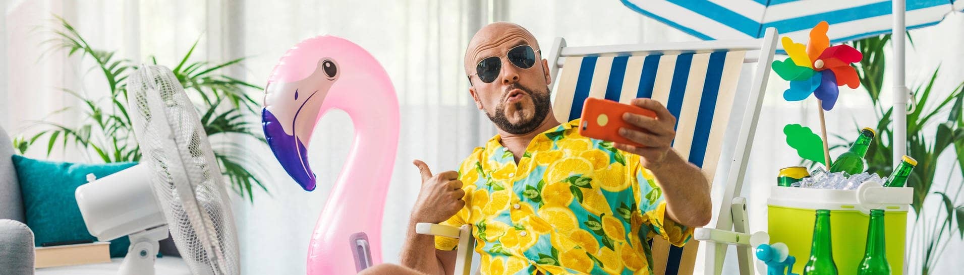 Junger Mann mit Sonnenbrille und buntem Hemd sitzt auf einem Liegestuhl zu Hause, das aussieht wie im Urlaub – neben ihm ein Ventilator und ein aufblasbarer Flamingo und ein Sonnenschirm. Er zeigt mit dem Daumen nach oben und hält ein Handy für das Urlaubsfoto vors Gesicht.