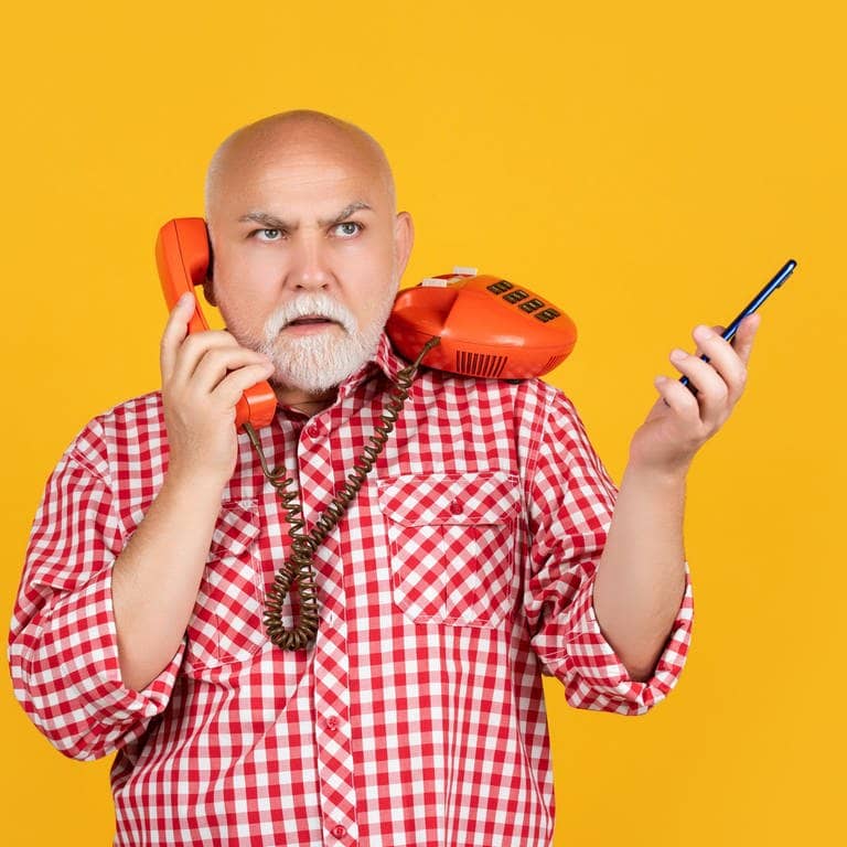 Ein Mann mit kariertem Hemd hält einen Hörer eines Festnetztelefons, das immer weniger genutzt wird, in der Hand. Das Gerät dazu steht auf der anderen Schulter.