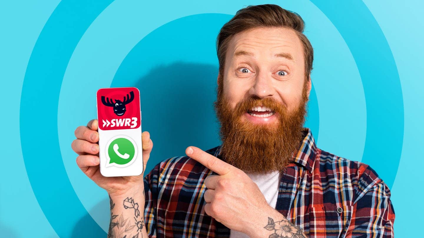 Ein Mann mit Bart hält vor blauem Hintergrund ein Handy in der Hand und zeigt darauf. Auf dem Bildschirm sind die Logos von SWR3 und Whatsapp zu sehen, da SWR3 nun auch einen Whatsapp-Kanal hat.