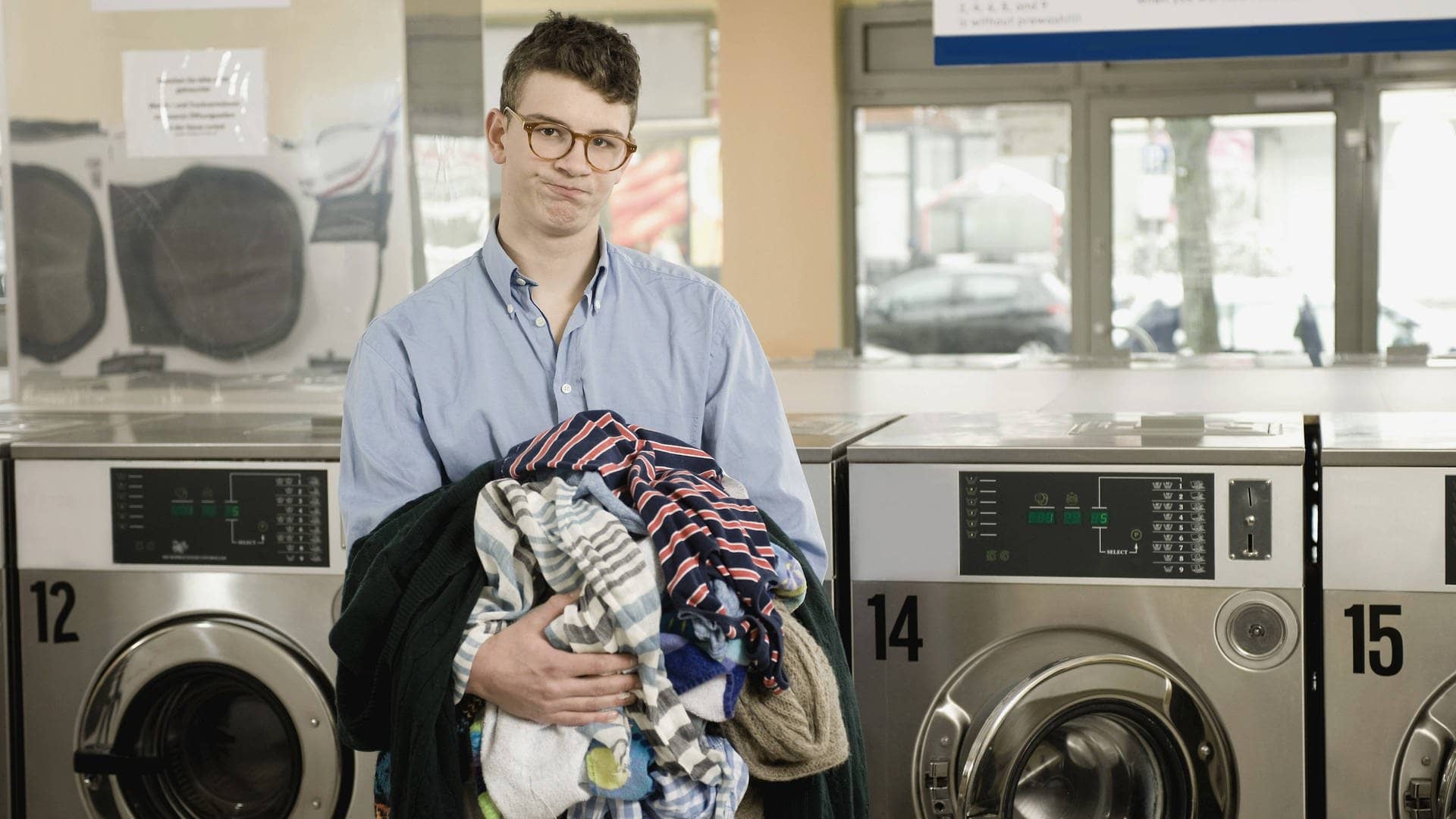 Mann schaut komisch aus der Wäsche, mit Wäsche in der Hand in einem Waschsalon