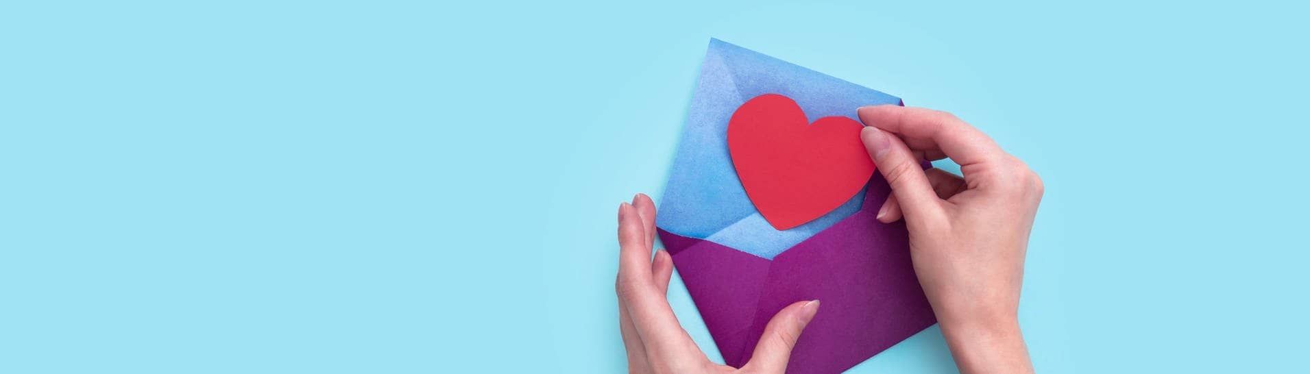 Eine Hand steckt ein rotes Papierherz in einen lila Umschlag.