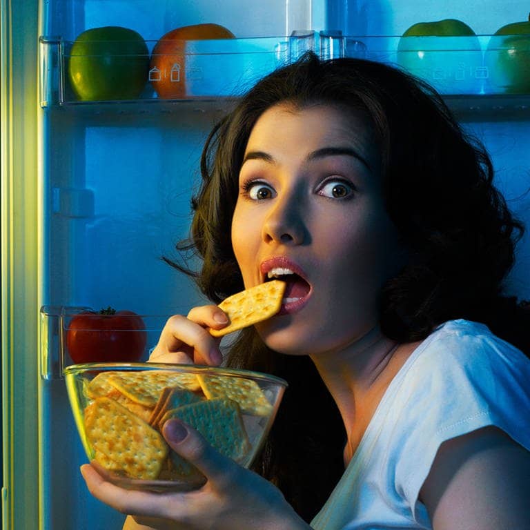 Frau isst salzigen Keks, vor offenem Kühlschrank 