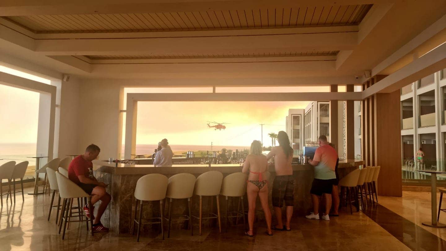 Menschen sitzen an einer Hotelbar, während im Hintergrund ein Hubschrauber vor dem Fenster vorbeifliegt.