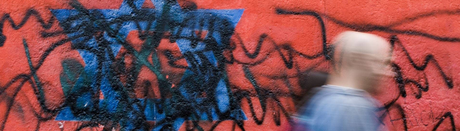 Berlin: Passanten gehen an dem zerstörten Wandbild des Künstlers Günther Schäfer an der East Side Gallery vorbei. Das Wandbild „Vaterland“, das die schwarz-rot-goldene Deutschlandfahne mit einem blauem Davidstern zeigt, wurde von Unbekannten mit schwarzen Graffitis beschmiert.