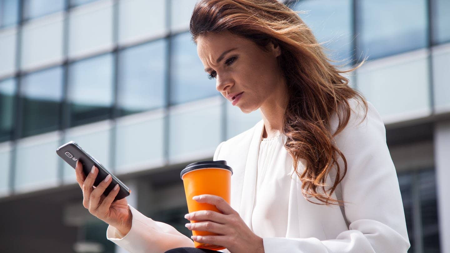 Eine junge Frau mit einem Kaffee in der Hand sitzt am Buerogebaeude auf einer Treppe und schaut irritiert auf ihr Smartphone.