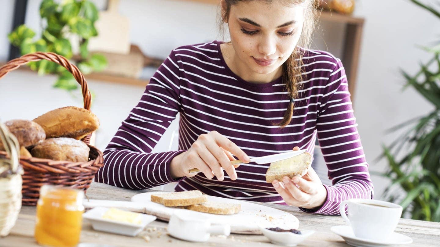 Junge Frau sitzt in der Küche am Frühstückstisch und schmiert sich ein Brot mit Butter