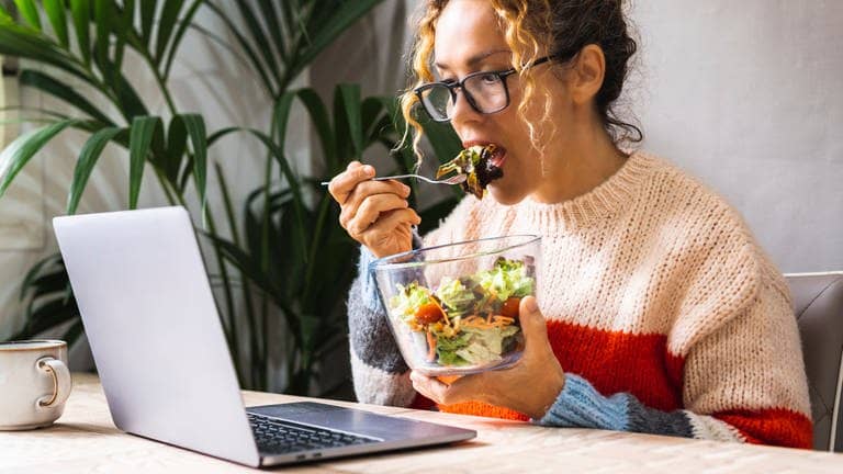 Junge Frau mit Brille sitzt am Esstisch vor dem Arbeits-Rechner. Sie macht eine Pause von der Arbeit, so wie es im Gesetz verordnet ist, schaut aber konzentriert auf den Laptop, als ob sie weiterhin arbeitet. Sie isst einen Salat.