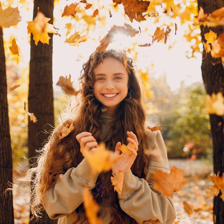 Herbstbild einer jungen Frau, die im Wald und im Sonnenschein in die Kamera lächelt. Um sie herum fallen Herbstblätter auf den Boden. Symbolbild für die vielen Gründe, warum der November eingentlich ein ganz toller Monat ist. 