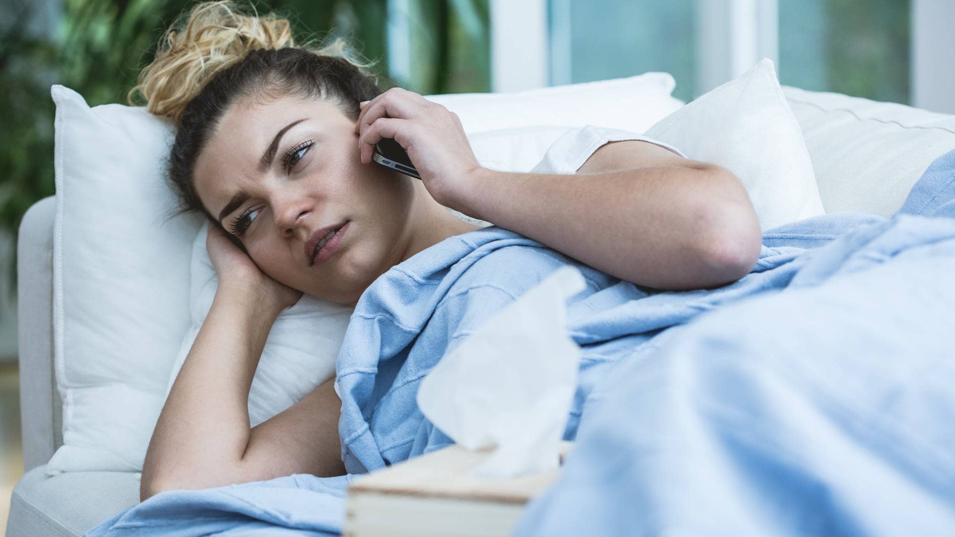 Die telefonische Krankmeldung beim Arbeitgeber macht es für so manchen Arbeitnehmer einfacher: Hier liegt eine Frau im Bett mit Telefon am Ohr und Taschentüchern auf dem Bett.