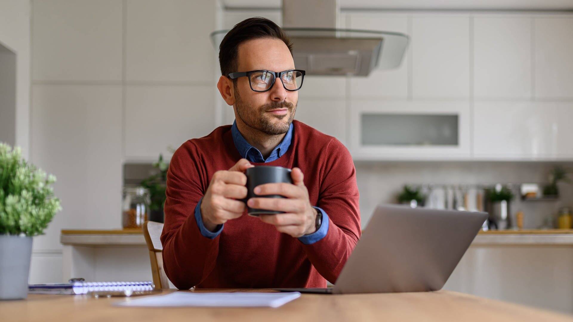 Mann mit Kaffeetasse auf dem Tisch sitzt in der Küche vor einem Laptop. Er denkt über eine mögliche Beförderung im Job nach und sucht nach Tipps für den Aufsteig und die Karriere.