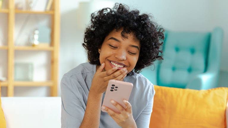 Junge Frau mit schwarzer Hautfarbe schaut auf ihr Smartphone und lacht – über kuriose Redewendungen aus anderen Sprachen.