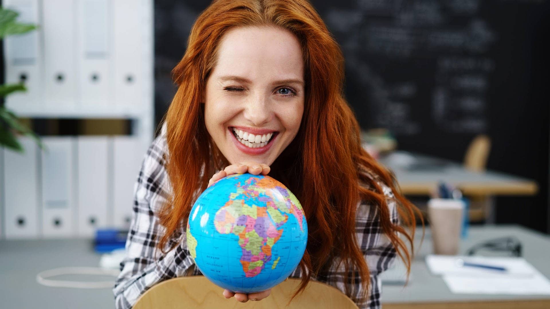Junge Frau in einem Klassenzimmer hält einen Globus in der Hand. Sie zwinkert und freut sich bestimmt über lustige Wörter aus anderen Sprachen.