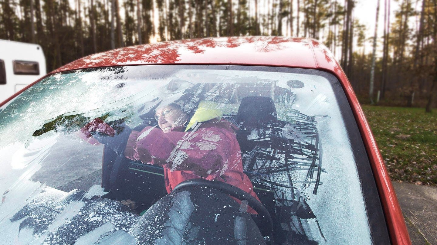 Die Innenscheibe im Auto ist gefroren und vereist. Eine Frau, dick eingepackt in Winterklamitten, kratzt die Scheibe von innen frei.