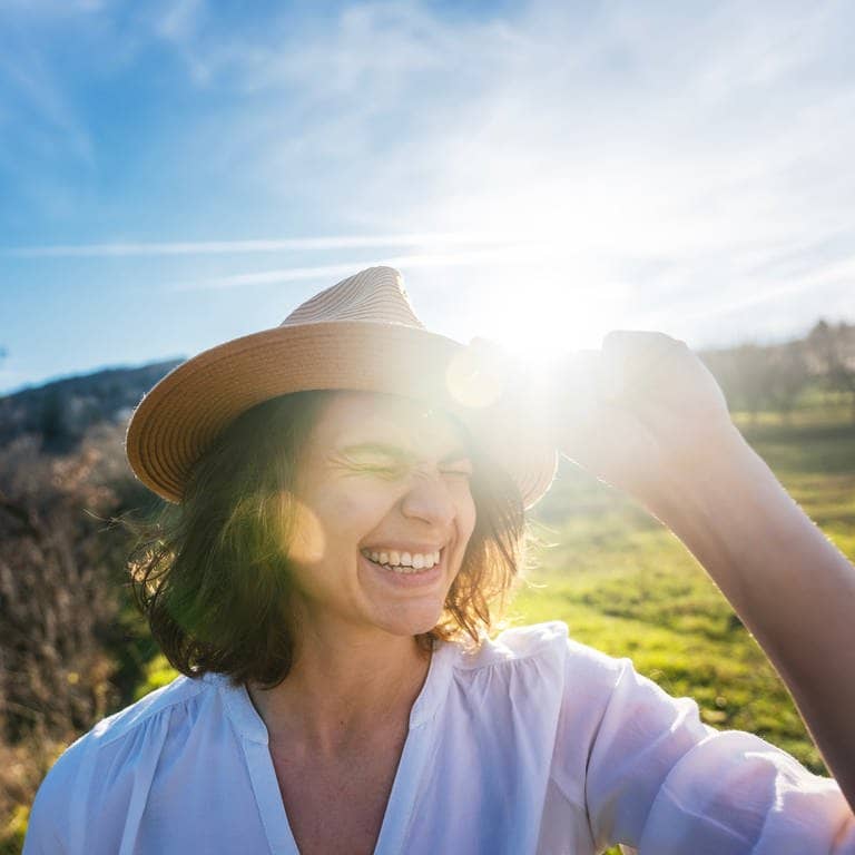 Eine junge Frau mit Hut und Hemd lacht in die Kamera, im Hintergrund kommt in einer Frühlingslandschaft die Sonne durch. Das Wetter wird jetzt endlich besser, erklärt SWR3-Wetterfrau Lea Spindler. (Foto: Adobe Stock, olezzo)