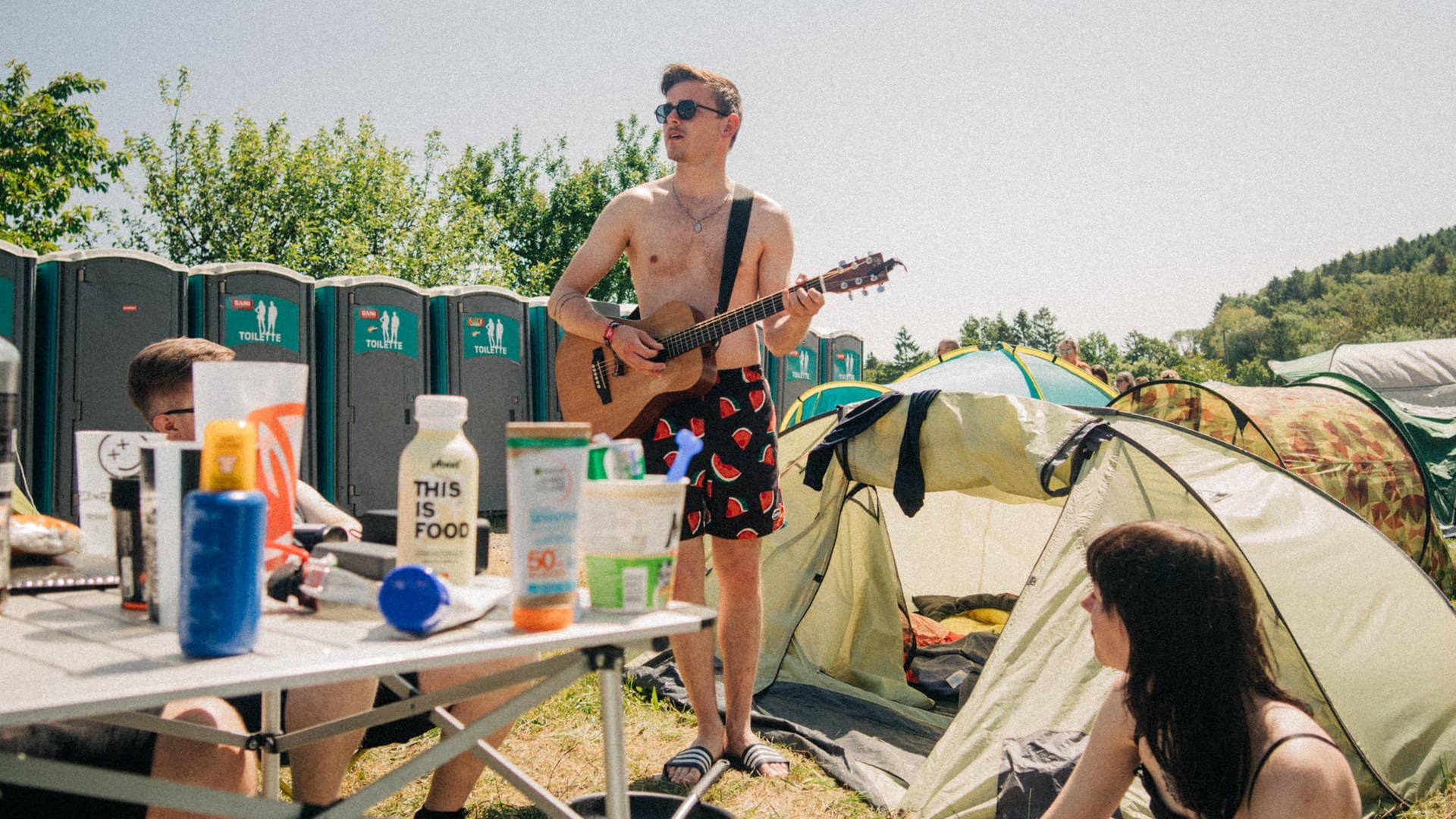 Ein junger Mann steht in Shorts und mit Gitarre auf dem Campingplatz von Rock am Ring zwischen Zelten