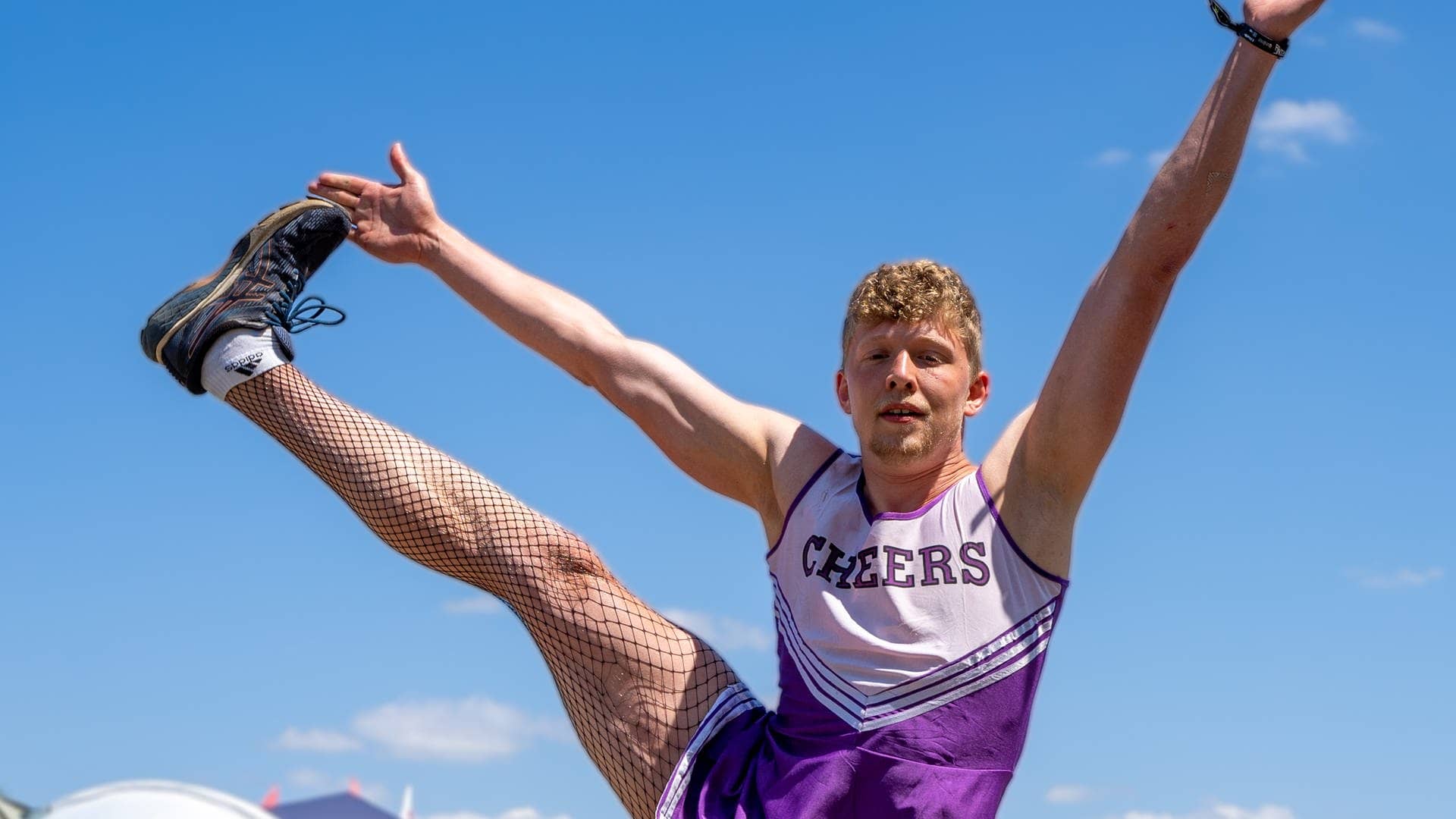 Ein junger Mann hat ein lila Cheerleader-Köstüm an und streckt sein Bein seitlich in die Höhe