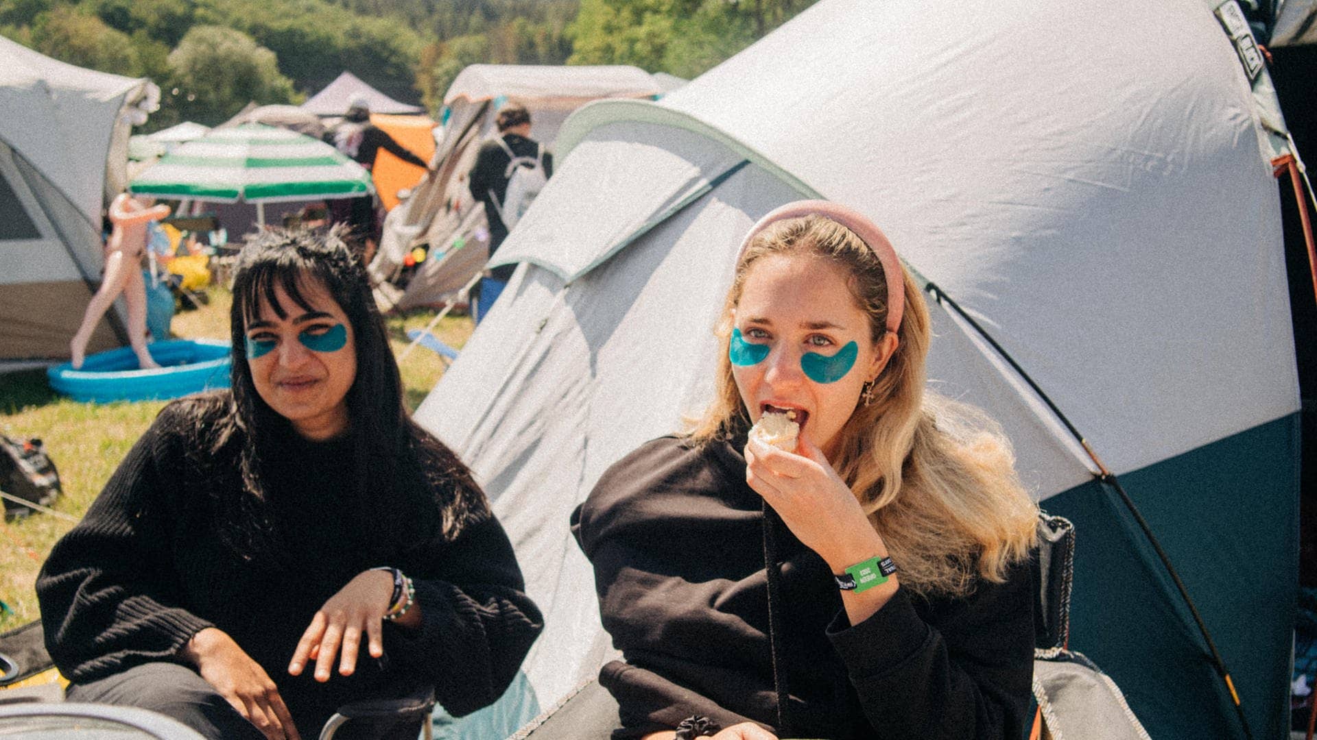 Zwei junge Frauen sitzen zwischen Zelten bei Rock am Ring mit Augenpads am Campintisch und frühstücken