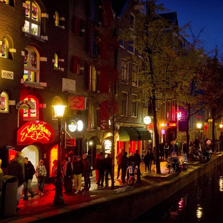 Menschen schlendern in Amsterdam am Abend an einer Gracht entlang durch den Rotlichtbezirk De Wallen.