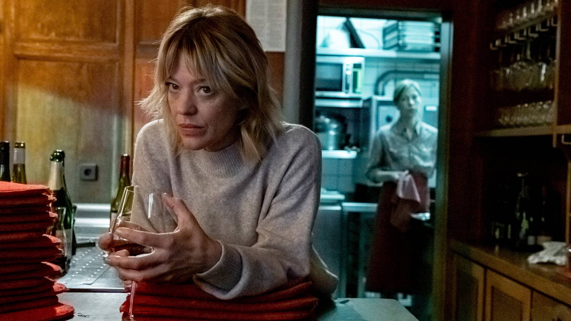 Tatort-Kritik Mainz „Aus dem Dunkel“: Ellen Berlinger mit einem Glas Wein hinter einer Theke. Im Hintergrund steht Maja in der Küche.