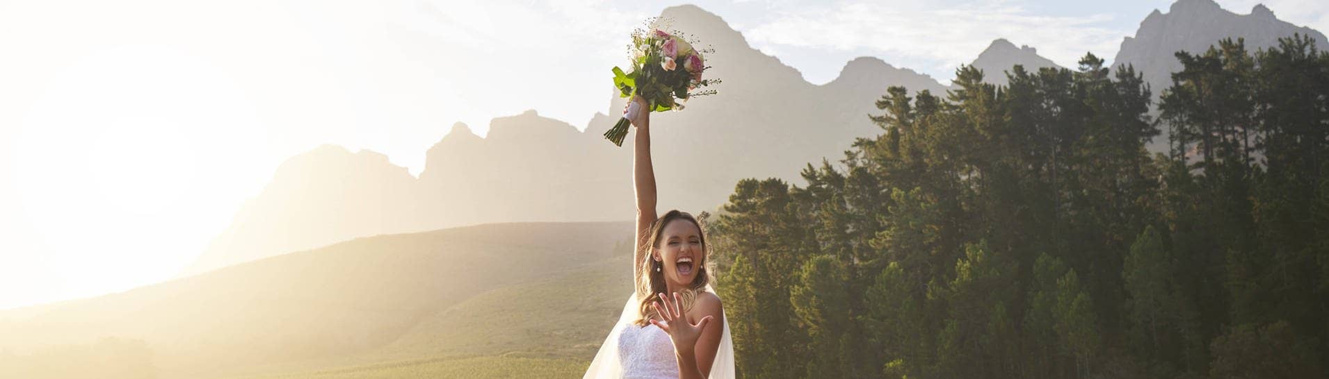 Eine Braut steht bei ihrer Hochzeit auf einem Steg an einem See und hält ihren Brautstrauß in die Höhe. Sie freut sich, weil sie bei ihrer Hochzeit mit günstigen DIY-Tipps Geld sparen konnte.