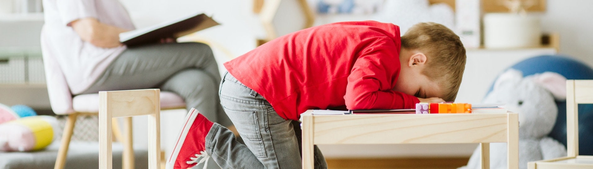 Kleiner Junge sitzt verzweifelt an einem Tisch. Frau im Hintergrund ist seine Therapeutin. (Foto: Adobe Stock/Photografee.eu)