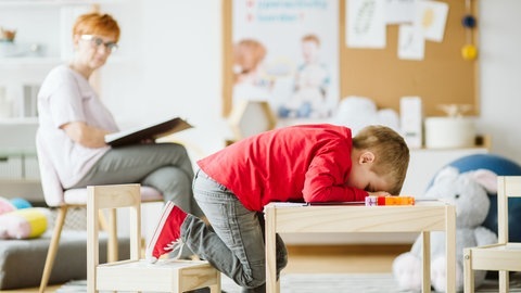 Kleiner Junge sitzt verzweifelt an einem Tisch. Frau im Hintergrund ist seine Therapeutin. (Foto: Adobe Stock/Photografee.eu)