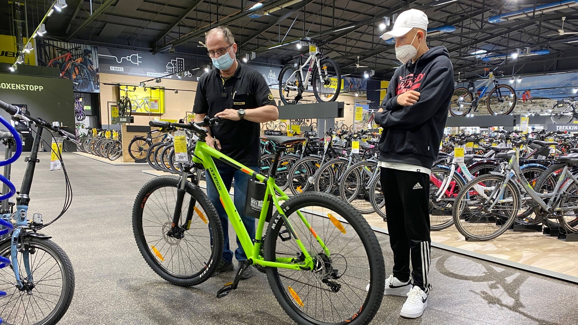 Foto: IMAGO, IMAGO / Sven Simon Ein Teenager beim Fahhradkauf in einem Zweirad Fachhandel. Ein Verkauefer beraet den jungen Kunden und stellt den Lenker eines grünen Bikes ein. (Foto: IMAGO, IMAGO / Sven Simon)