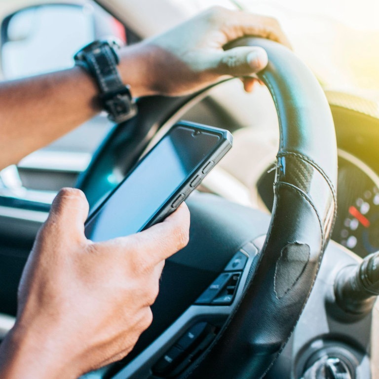 Mann hält Telefon am Steuer in der Hand (Foto: IMAGO, imagebroker)