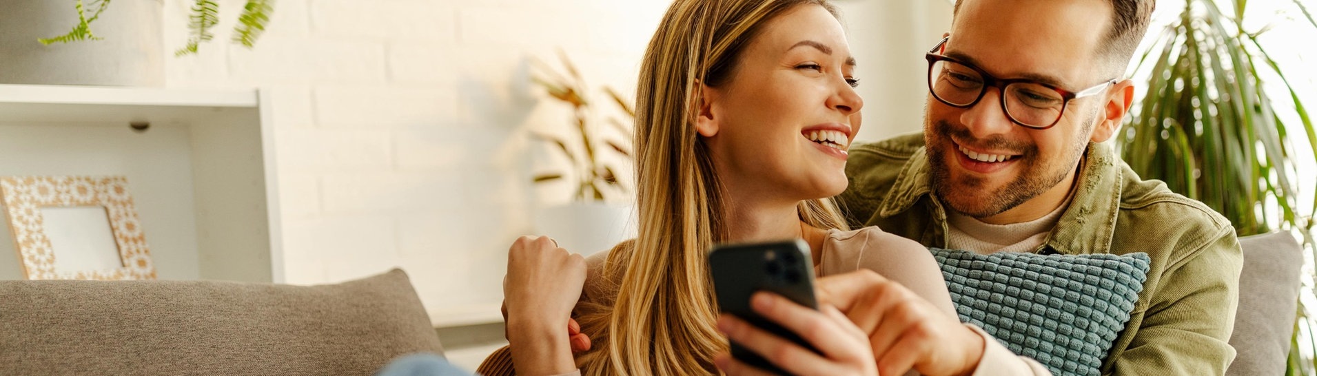 Junges Paar sitzt auf der Couch, liest auf dem Smartphone, lacht und freut sich über gute Nachrichten. (Foto: Adobe Stock, Adobe Stock/Nebojsa)