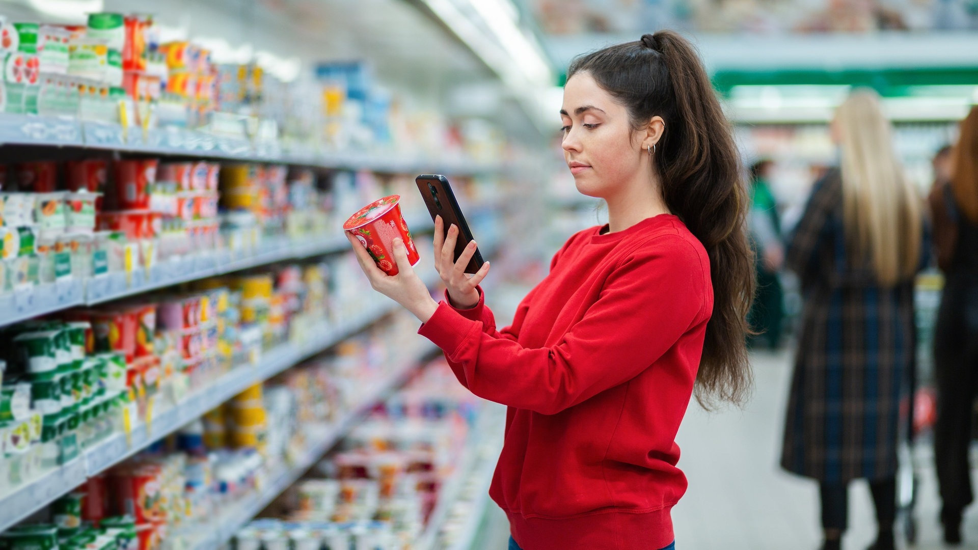 Foto: Adobe Stock, _KUBE_ Eine junge Frau scannt den QR-Code auf einer Joghurtpackung. Im Hintergrund ein Supermarkt mit verschwommenen Besuchern. Das Konzept moderner Technologien, Nutzt sie eine Spar-App? (Foto: Adobe Stock, _KUBE_)
