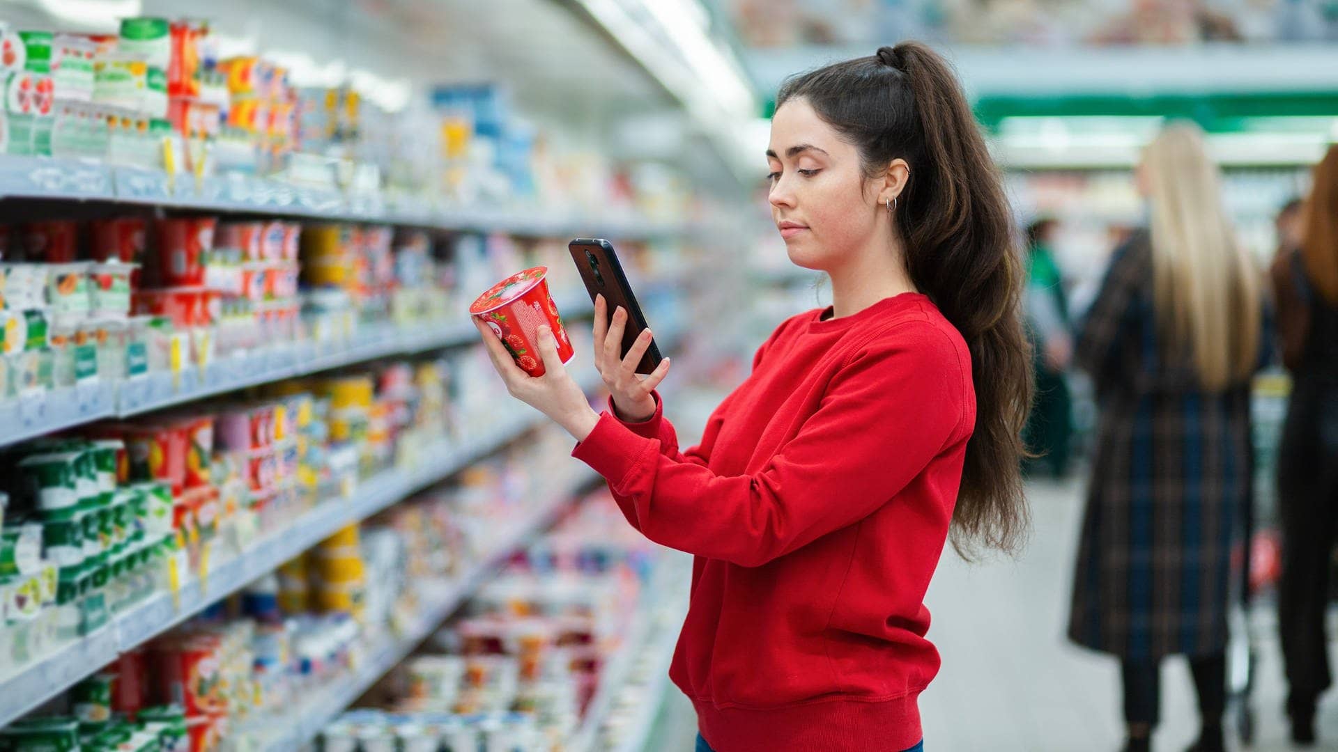 Eine junge Frau scannt den QR-Code auf einer Joghurtpackung. Im Hintergrund ein Supermarkt mit verschwommenen Besuchern. Das Konzept moderner Technologien, Nutzt sie eine Spar-App? (Foto: Adobe Stock, _KUBE_)