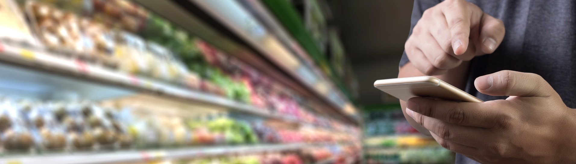 Männliche Hände tippen auf einem handy rum. Im Hintergrund die Kühltheke eines Supermarkts (Foto: Adobe Stock, onephoto)