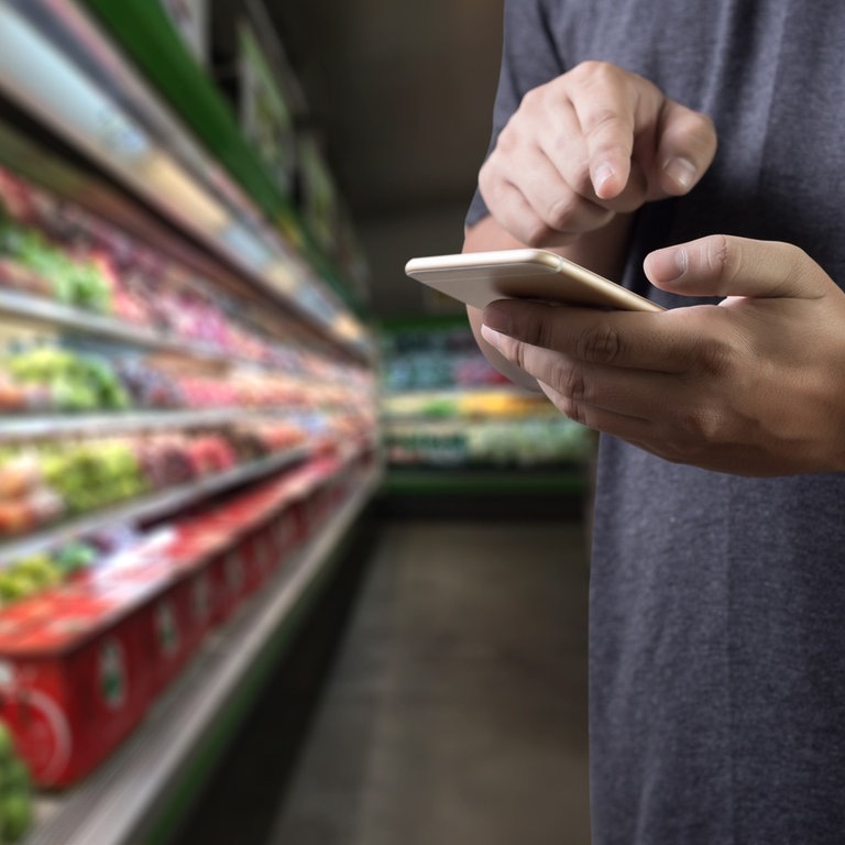 Männliche Hände tippen auf einem handy rum. Im Hintergrund die Kühltheke eines Supermarkts (Foto: Adobe Stock, onephoto)