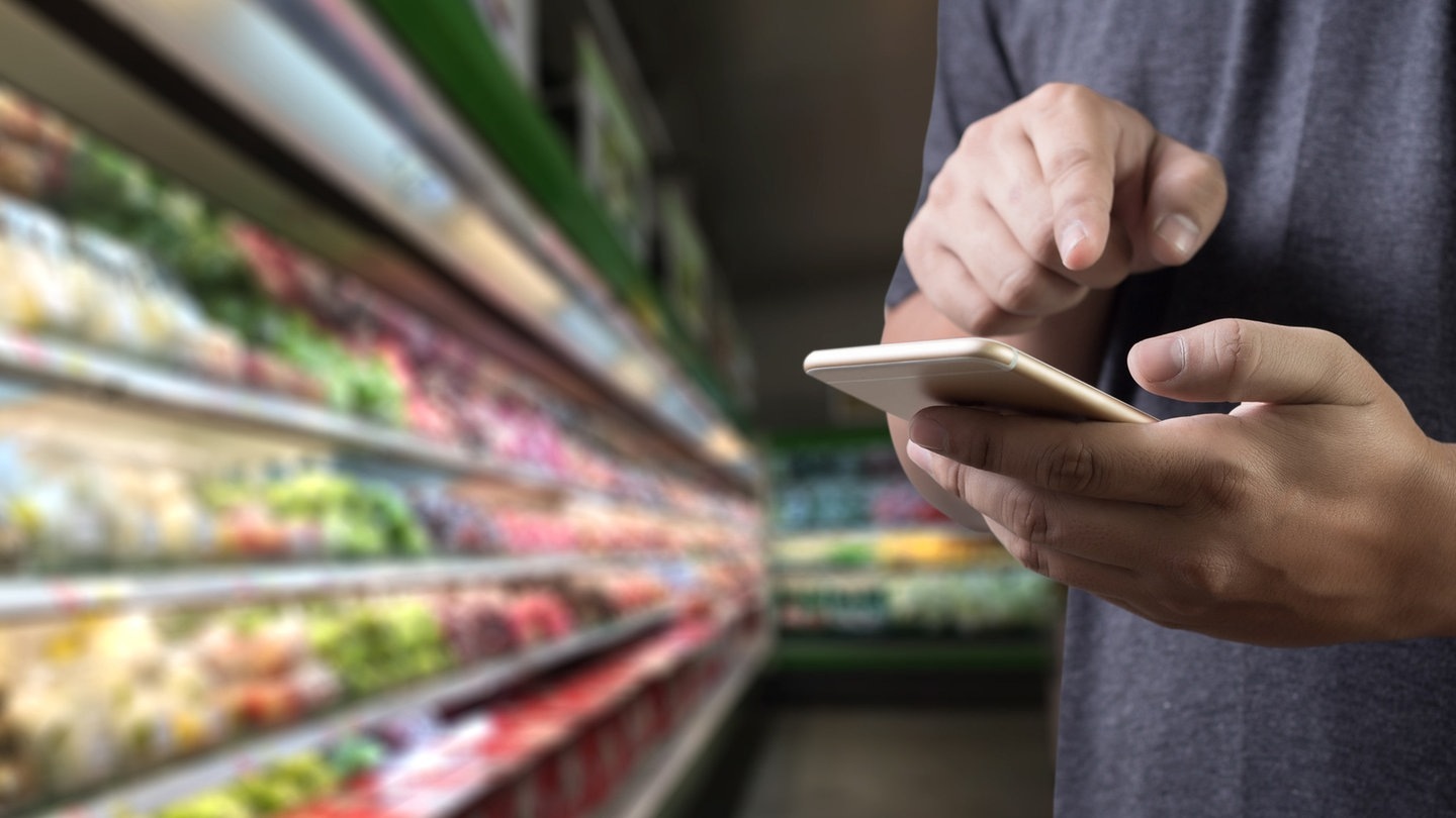 Männliche Hände tippen auf einem handy rum. Im Hintergrund die Kühltheke eines Supermarkts (Foto: Adobe Stock, onephoto)