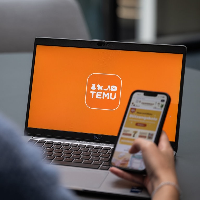 Temu App aus China: Erfahrungsberichte. Person hält Handy mit Temu App vor dem Laptop, auf dem der Onlineshop offen ist (Foto: picture-alliance / Reportdienste, picture alliance/dpa | Hannes P Albert)