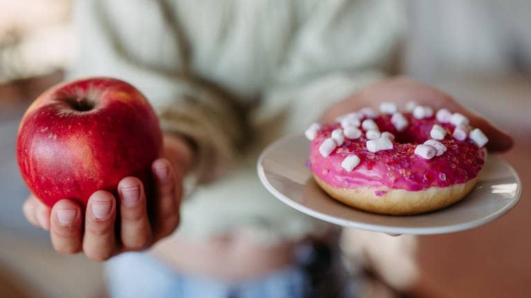 Frau hält einen Apfel und eine Donut. Beide enthalten Zucker und werden den Glukosespiegel ansteigen lassen