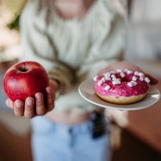 Frau hält einen Apfel und eine Donut. Beide enthalten Zucker und werden den Glukosespiegel ansteigen lassen