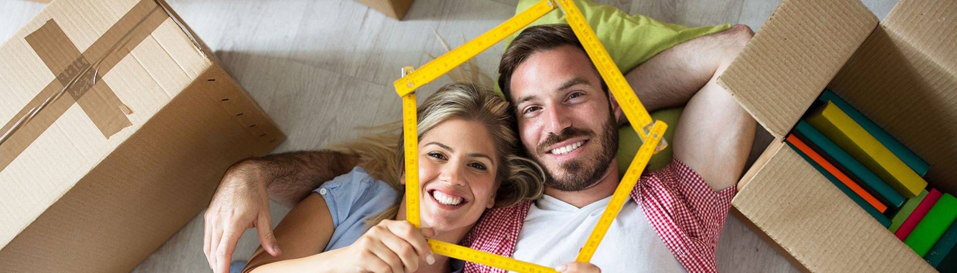 Mann und Frau liegen auf dem Boden, zwoschen Umzugskisten und lächeln in die Kamera  (Foto: Adobe Stock / didesign)