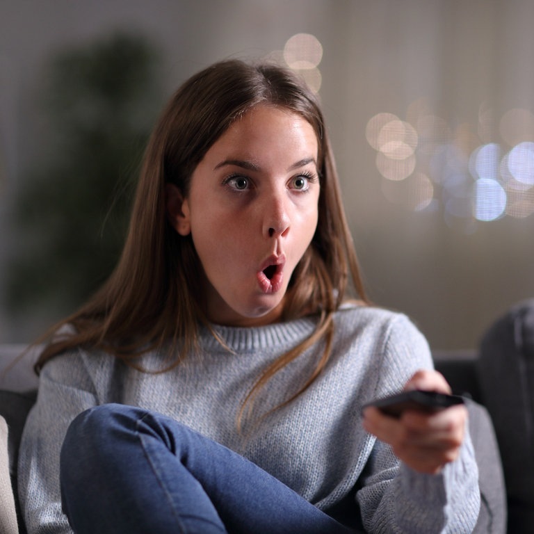 Überrraschte Frau beim Frensehen zu Hause auf dem Sofa (Foto: Adobe Stock / Antonioguillem)