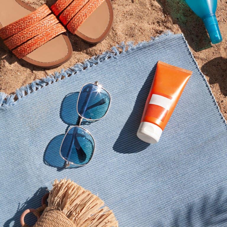 Sonnecremetube liegt auf Strandtuch mit Sonnenbrille und Sonnenschirm (Foto: Adobe Stock, mariarom)