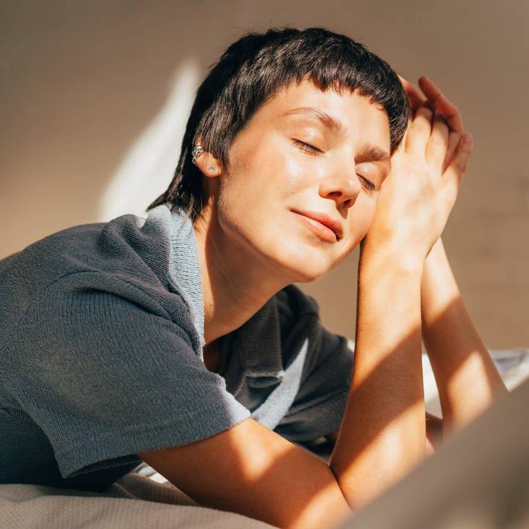 Frau liegt auf dem Bett im hellen Licht der Sonne, die durchs Fenster scheint (Foto: Adobe Stock, Ilona)