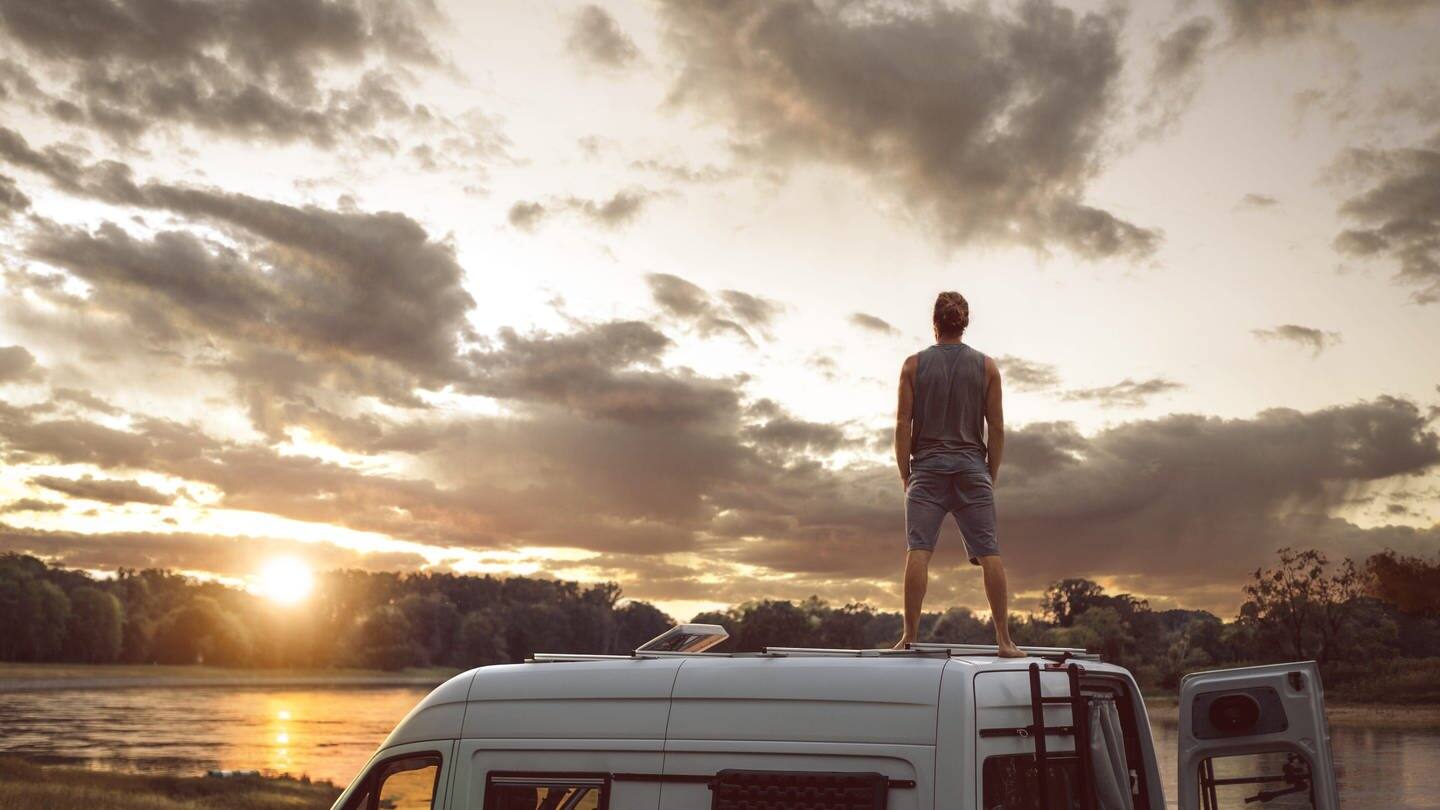 Mann steht auf dem Dach seines Wohnmobils und schaut in den Sonnenuntergang (Foto: Adobe Stock/photoschmidt)