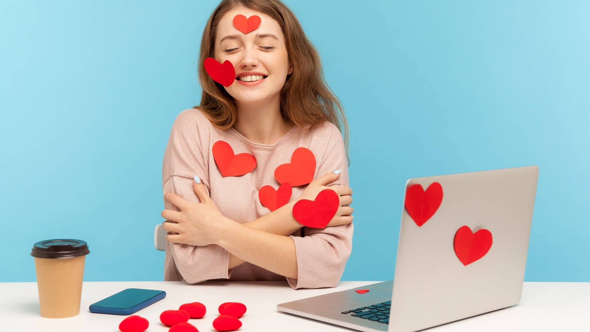 Junge Frau, beklebt mit Herzen, sitzt verliebt an einem Schreibtisch mit Laptop. (Foto: Adobe Stock/khosrork)
