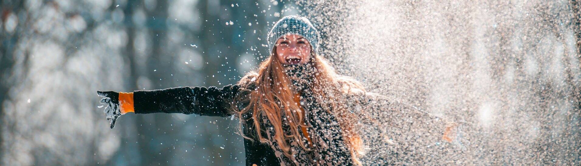 Junge Frau, warm angezogen mit Mütze, wirft Schnee in die Luft und freut sich (Foto: Adobe Stock/leszekglasner)