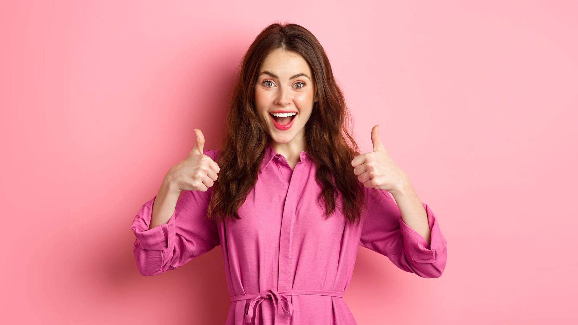 Junge Frau vor pinkfarbenem Hintergrund und mit pinkfarbener Bluse lacht und zeigt mit beiden Daumen in die Luft (Foto: Adobe Stock, Adobe Stock/Mix and Match Studio)