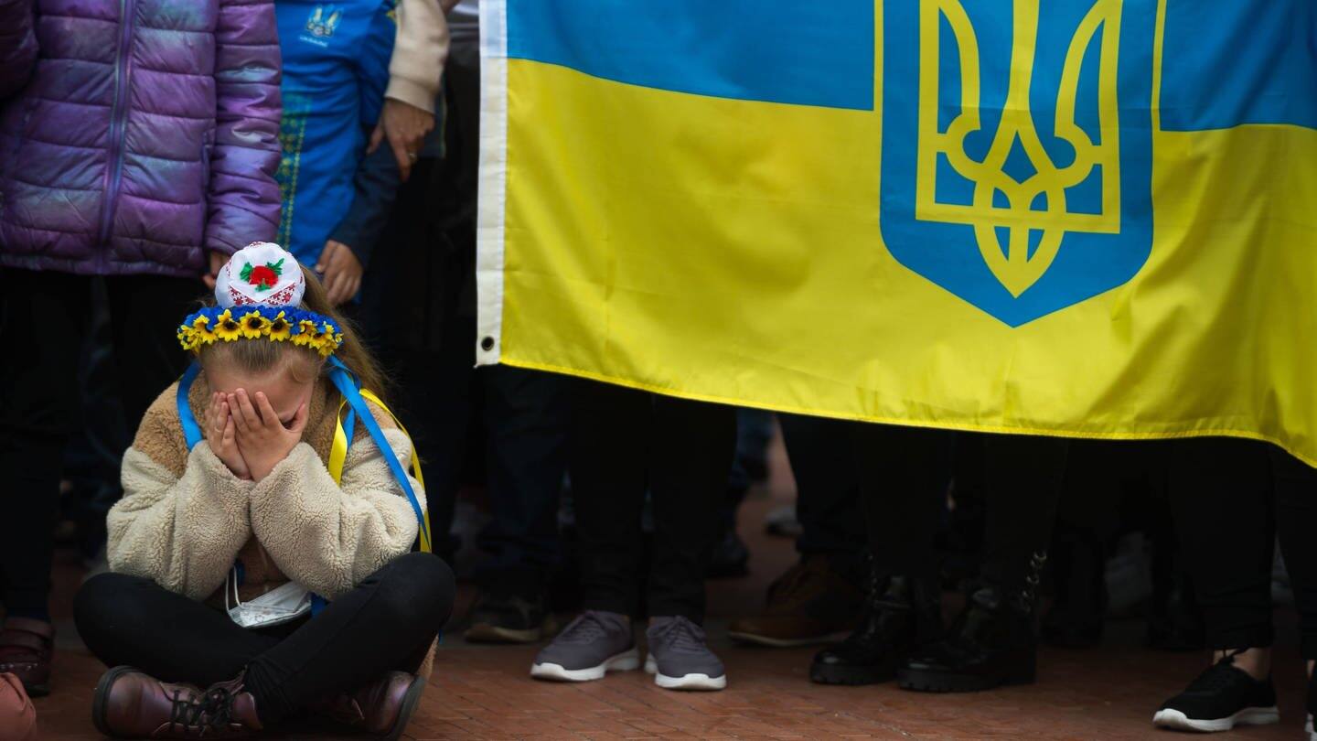 Ein kleines ukrainisches Mädchen sitzt auf dem Boden vor einer ukrainischen Flagge und schlägt die Hände vors Gesicht.  (Foto: IMAGO, IMAGO / ZUMA Wire)
