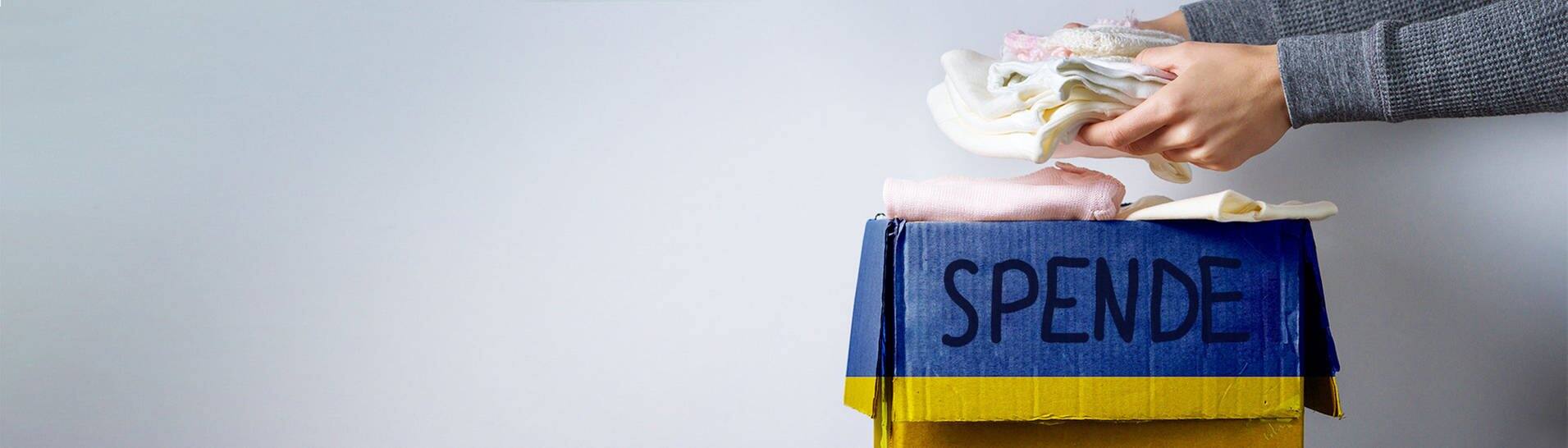 Karton mit der Aufschrift "Spende", jemand stapelt Kleidung hinein zur Hilfe für die Ukraine (Foto: Adobe Stock/Goffkein)