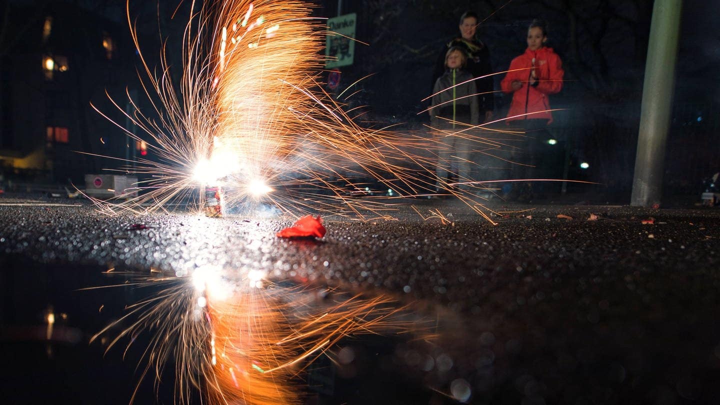 Mehrere Personen sehen einem funkensprühenden Bodenfeuerwerk zu. (Foto: IMAGO, imago/Marius Schwarz)