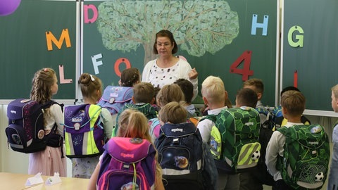 Schülerinnen und Schüler stehen mit ihren Schulränzen vor einer Lehrerin im Klassenraum. (Foto: dpa Bildfunk, picture alliance/dpa/dpa-Zentralbild | Matthias Bein) Schülerinnen und Schüler stehen mit ihren Schulränzen vor einer Lehrerin im Klassenraum.