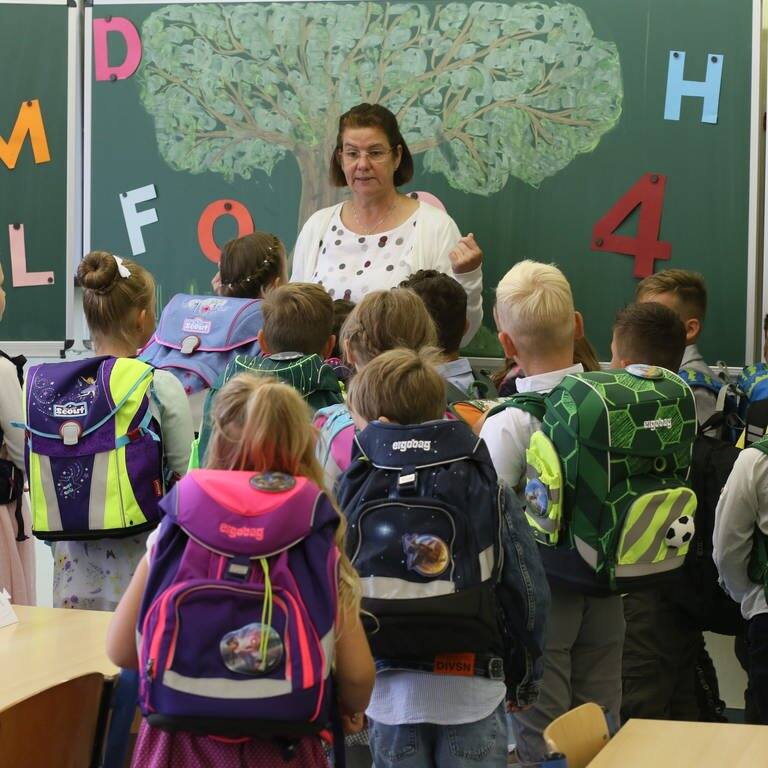 Schülerinnen und Schüler stehen mit ihren Schulränzen vor einer Lehrerin im Klassenraum.